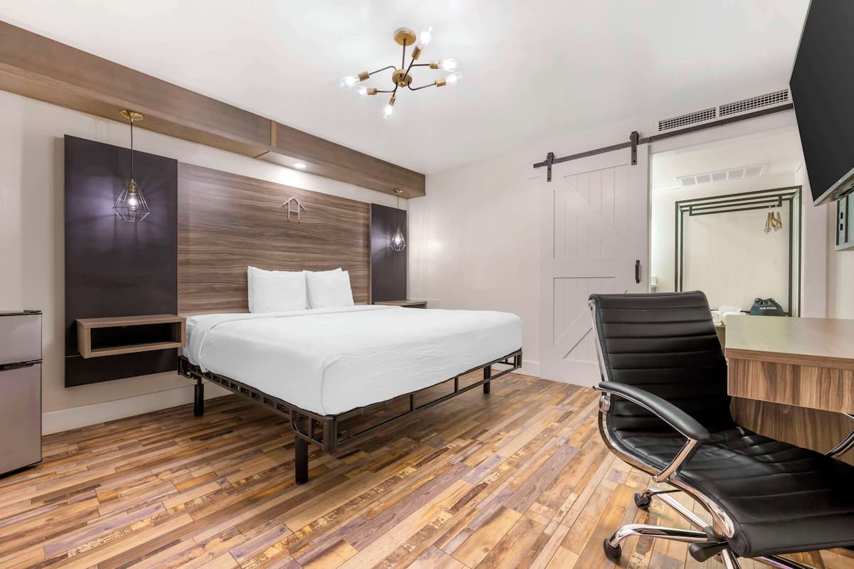 A modern bedroom with a wooden floor, a bed with white bedding, a black office chair, and a desk.