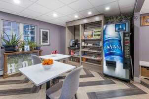 Um hall de hotel moderno com um balcão de recepção branco, uma máquina de café e um dispensador de água. Há plantas em vasos e uma pequena mesa com uma tigela de frutas.