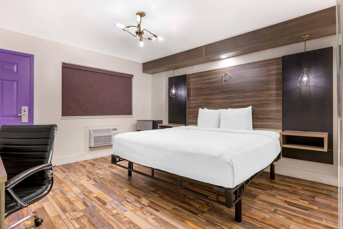 A bedroom with a wooden floor, a bed with white bedding, a black office chair, a purple door, and a modern ceiling light fixture.