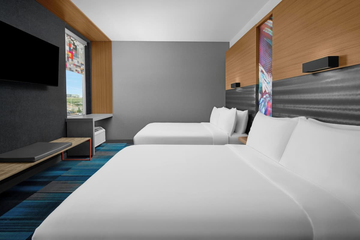 Una habitación de hotel moderna con dos camas, un televisor de pantalla plana montado en la pared, una mesita y un ventanal con vistas a la ciudad.