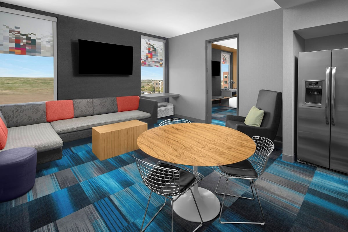 Habitación de hotel moderna con sofá, una mesa de comedor con sillas, televisor de pantalla plana, refrigerador y un ventanal con vistas a la ciudad.