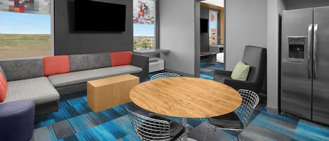 Een moderne hotelkamer met een bank, een eettafel met stoelen, een flatscreen televisie, een koelkast en een groot raam met uitzicht op de stad.
