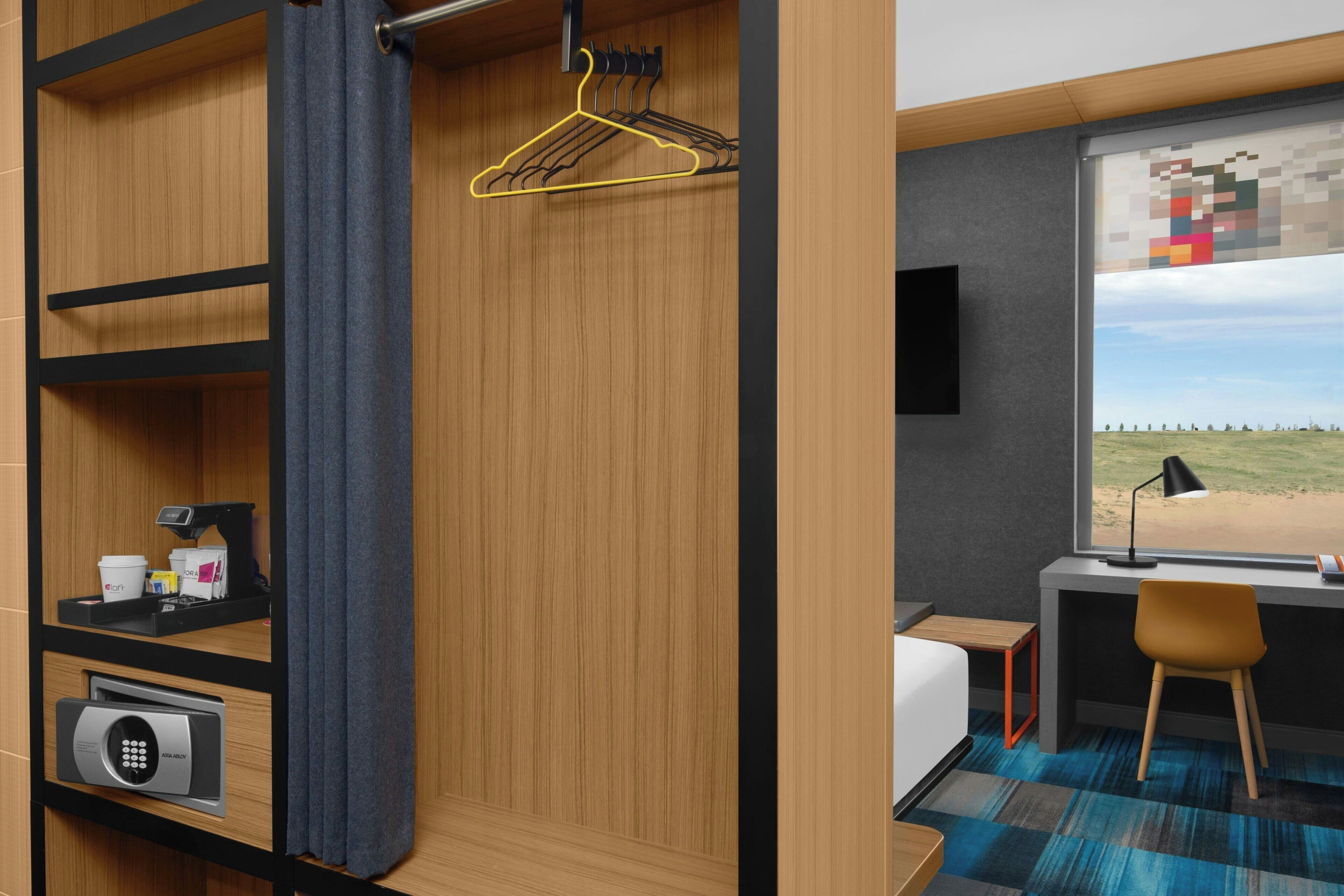 A compact room with a bed, a desk, a chair, a window with a view, a hanging clothes rack, and a security keypad.