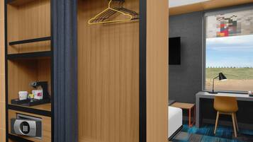 A compact room with a bed, a desk, a chair, a window with a view, a hanging clothes rack, and a security keypad.