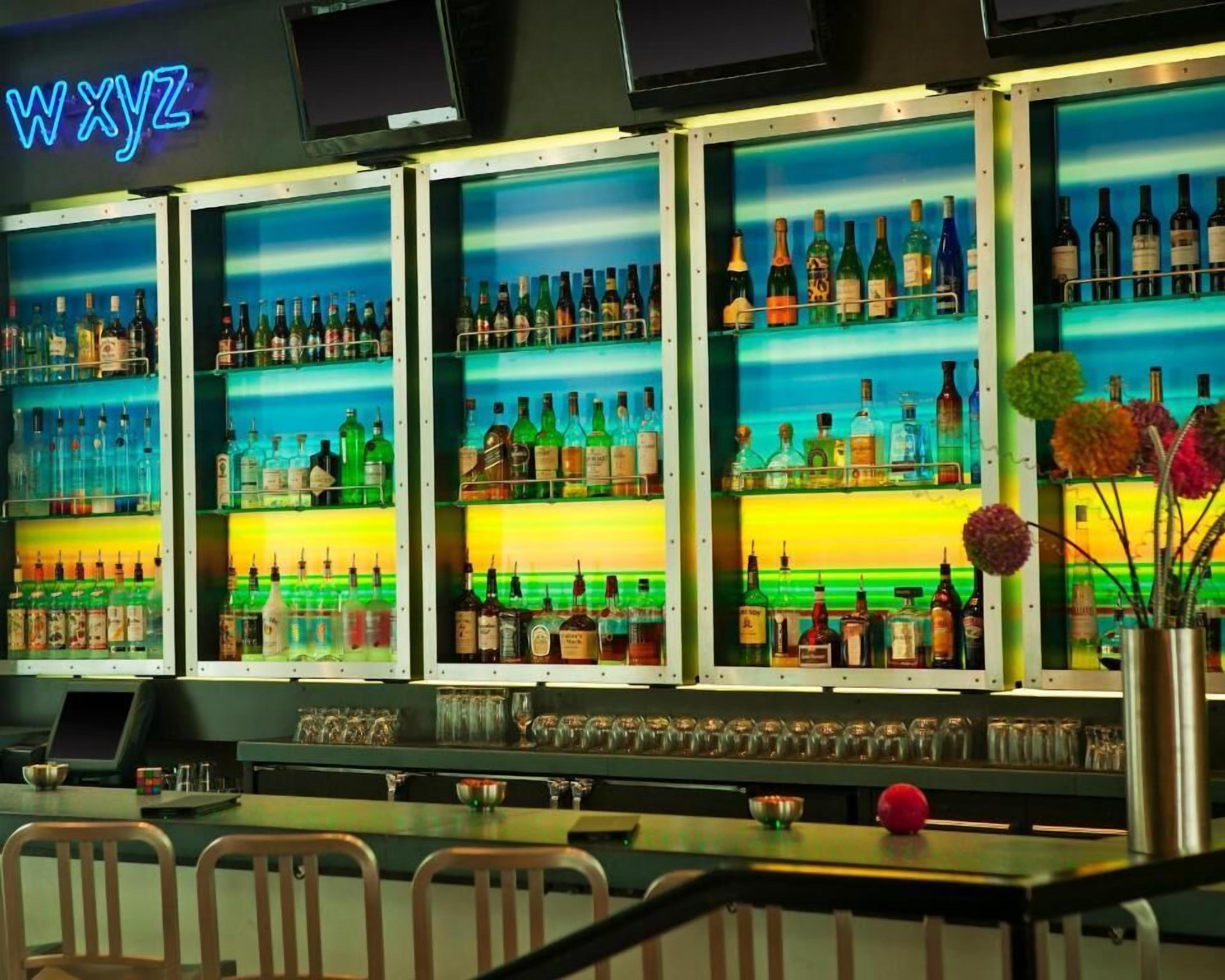 Eine Bar mit mehreren Regalen voller Spirituosenflaschen, einem Neonlicht und einer Bar, an der Stühle stehen.