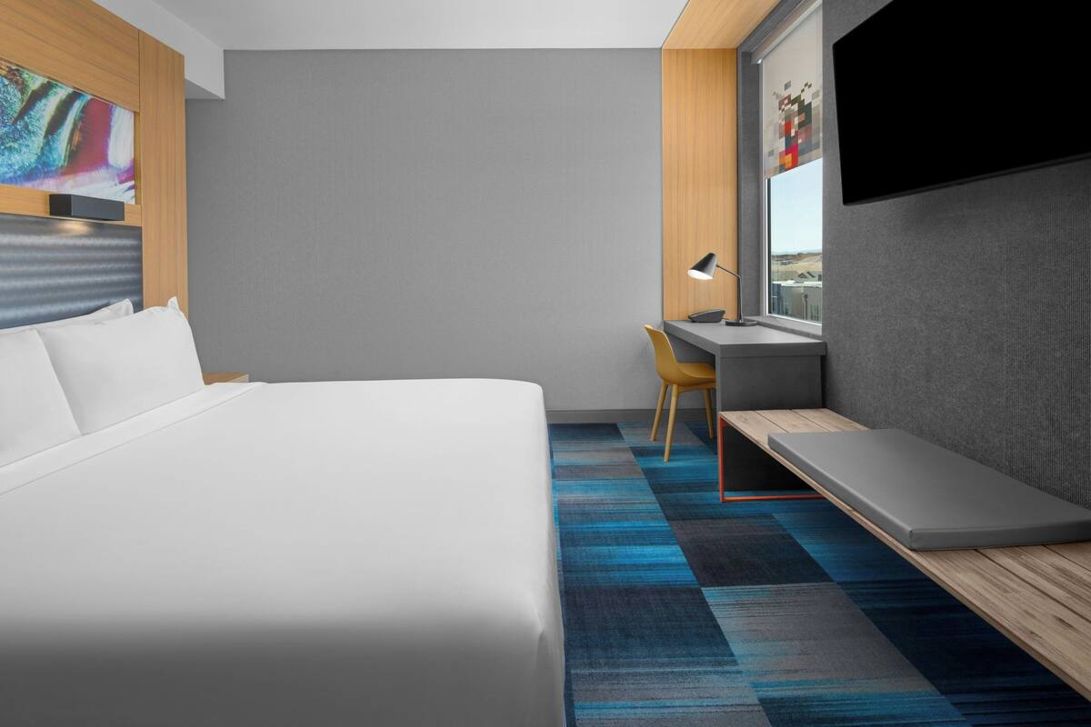 Una habitación de hotel moderna con una cama grande, un escritorio con una silla, un televisor montado en la pared y vistas a la ciudad por la ventana.