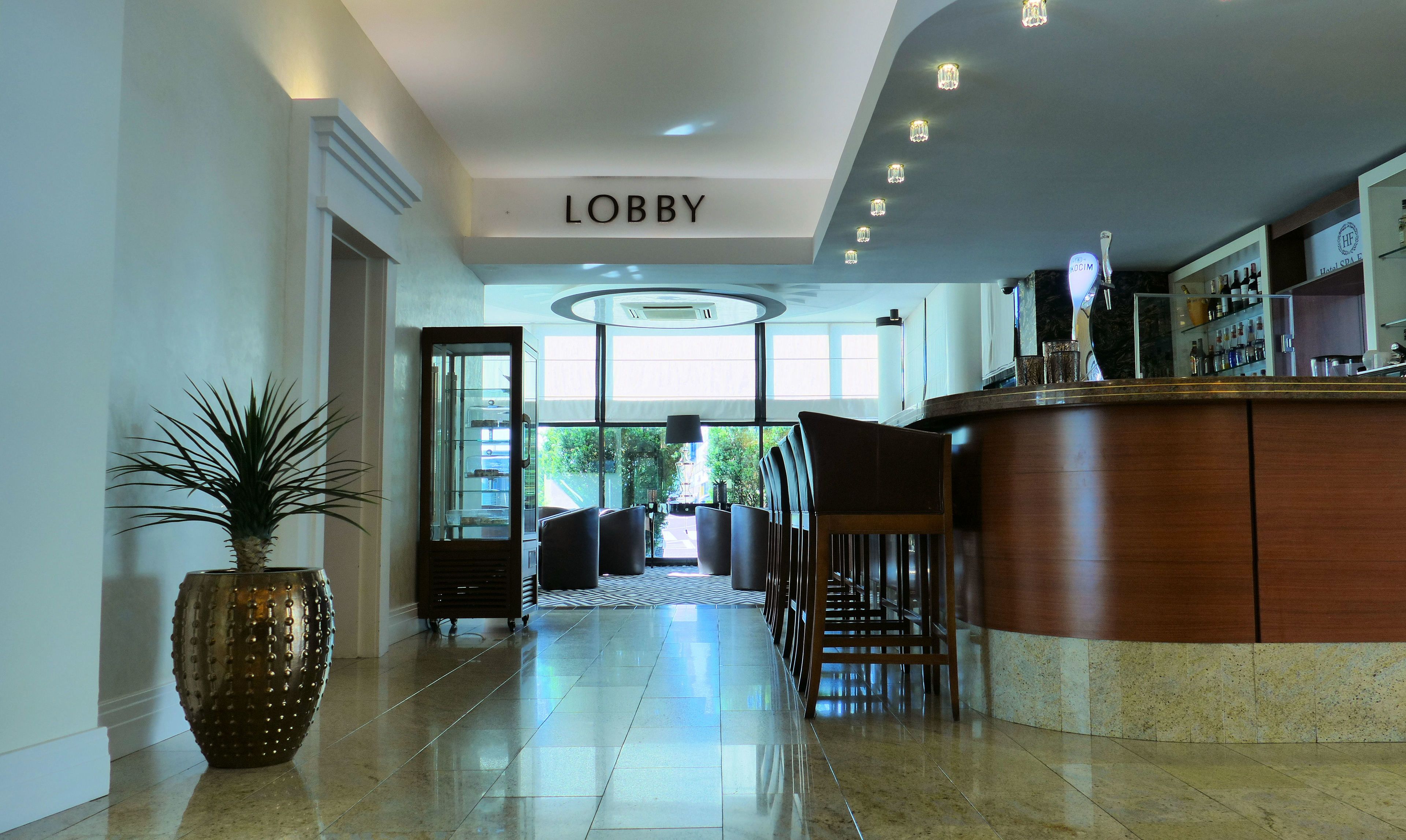 Un vestíbulo con un letrero que dice claramente “LOBBY”, una recepción, una planta grande en maceta y una zona de estar con sillas y un sofá.