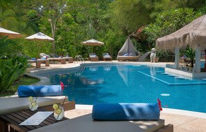 Zona de piscina con sillones de descanso, sombrillas y una estructura de techo de paja.