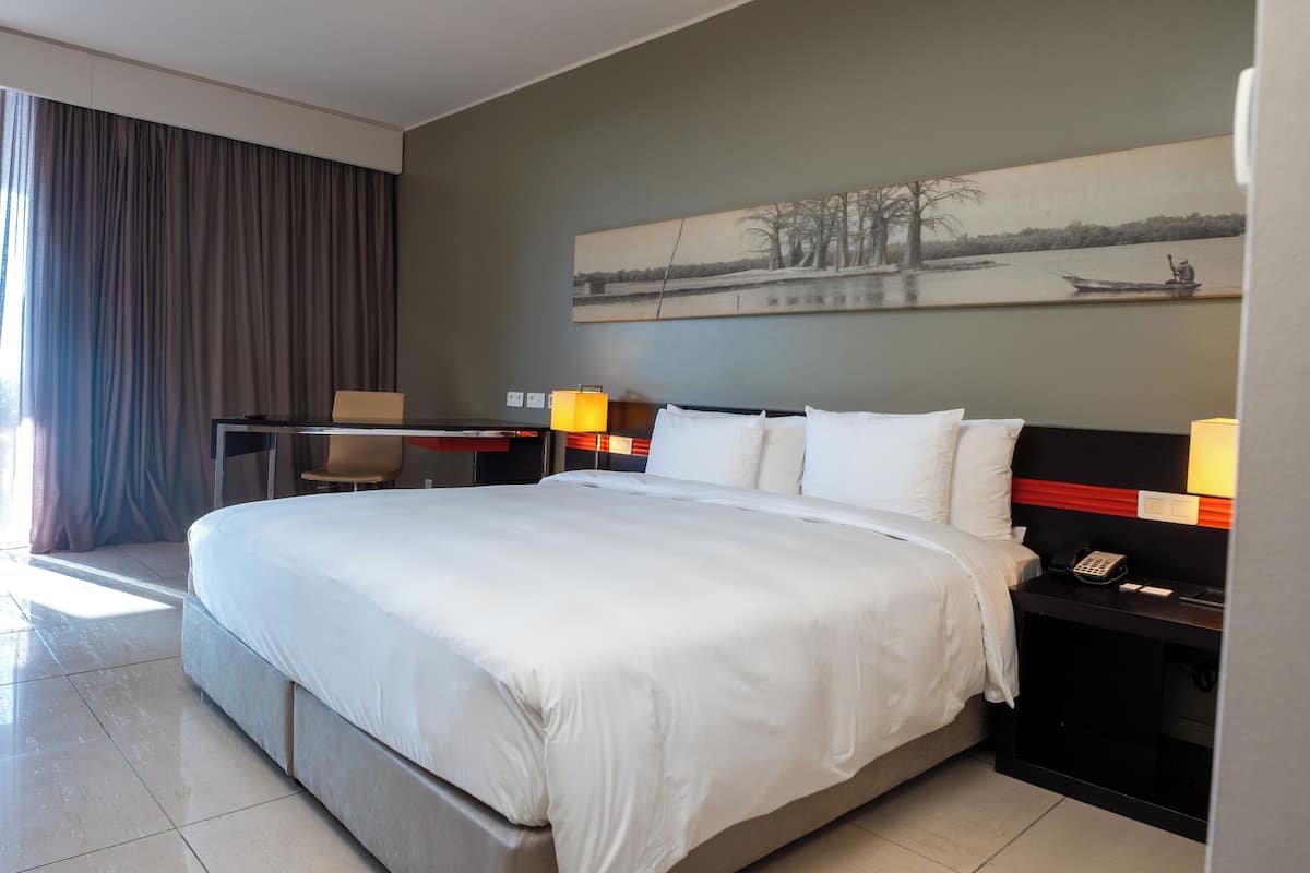 Ein modernes Hotelzimmer mit einem großen Bett, einem Schreibtisch mit Stuhl, einem Nachttisch mit Lampe und einem Bild an der Wand.
