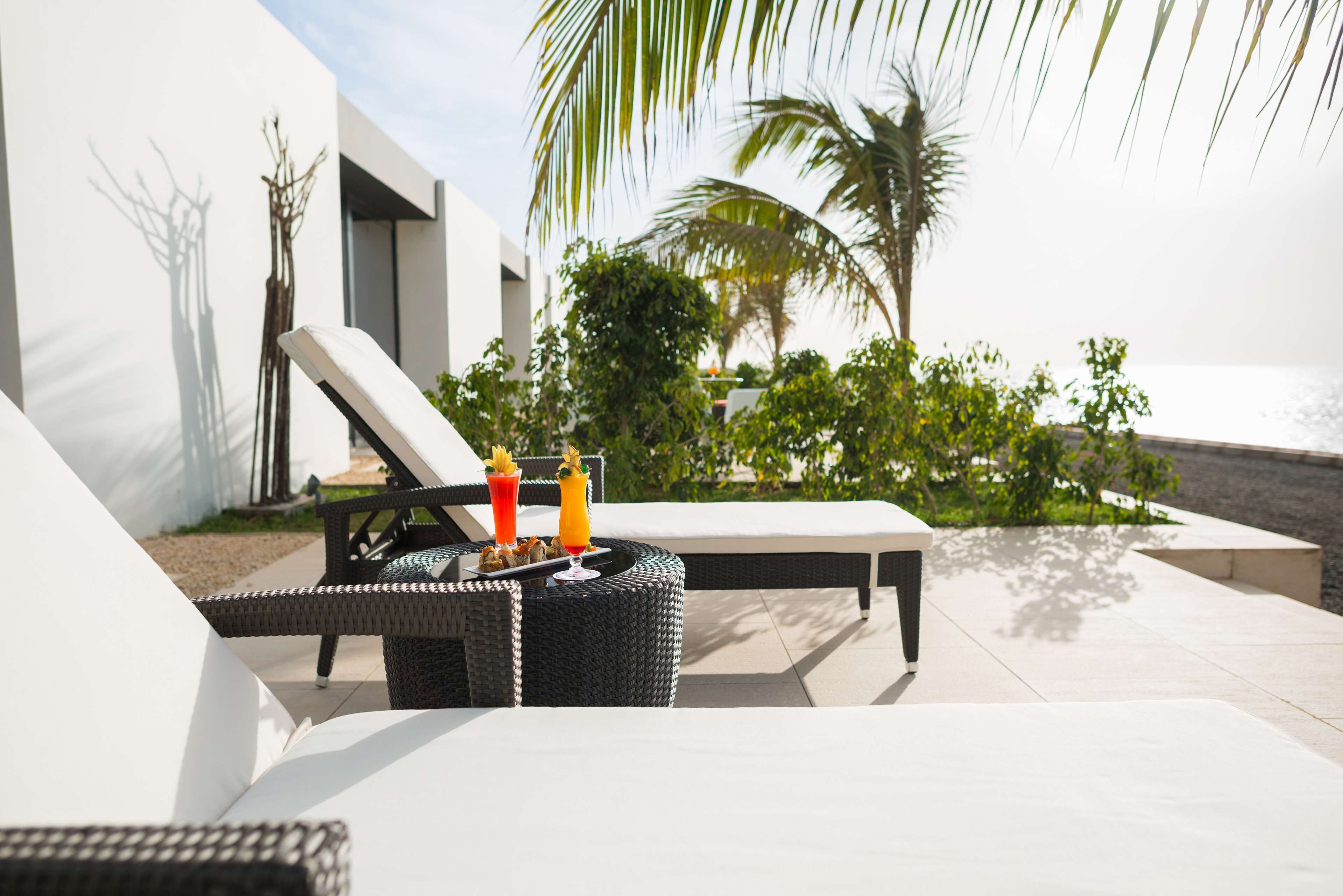 A sunlit outdoor lounge area with a wicker table, two lounge chairs, and a drink tray.