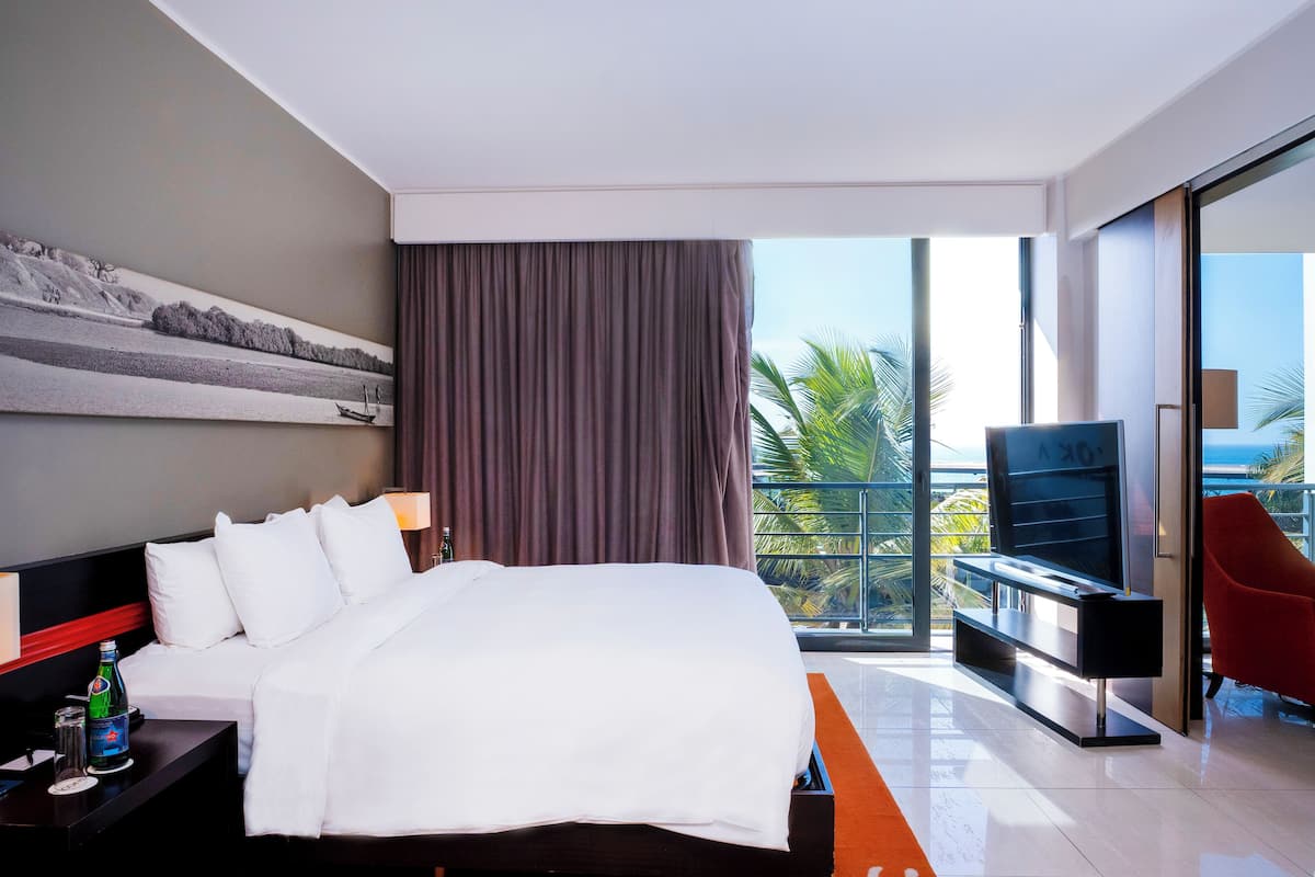 Ein modernes Hotelzimmer mit einem großen Bett, einem Flachbildfernseher, einem Balkon mit einem roten Sessel und Meerblick.