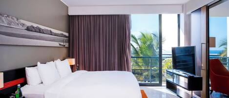 A modern hotel room with a large bed, a flat-screen TV, a balcony with a red chair, and a view of the ocean.