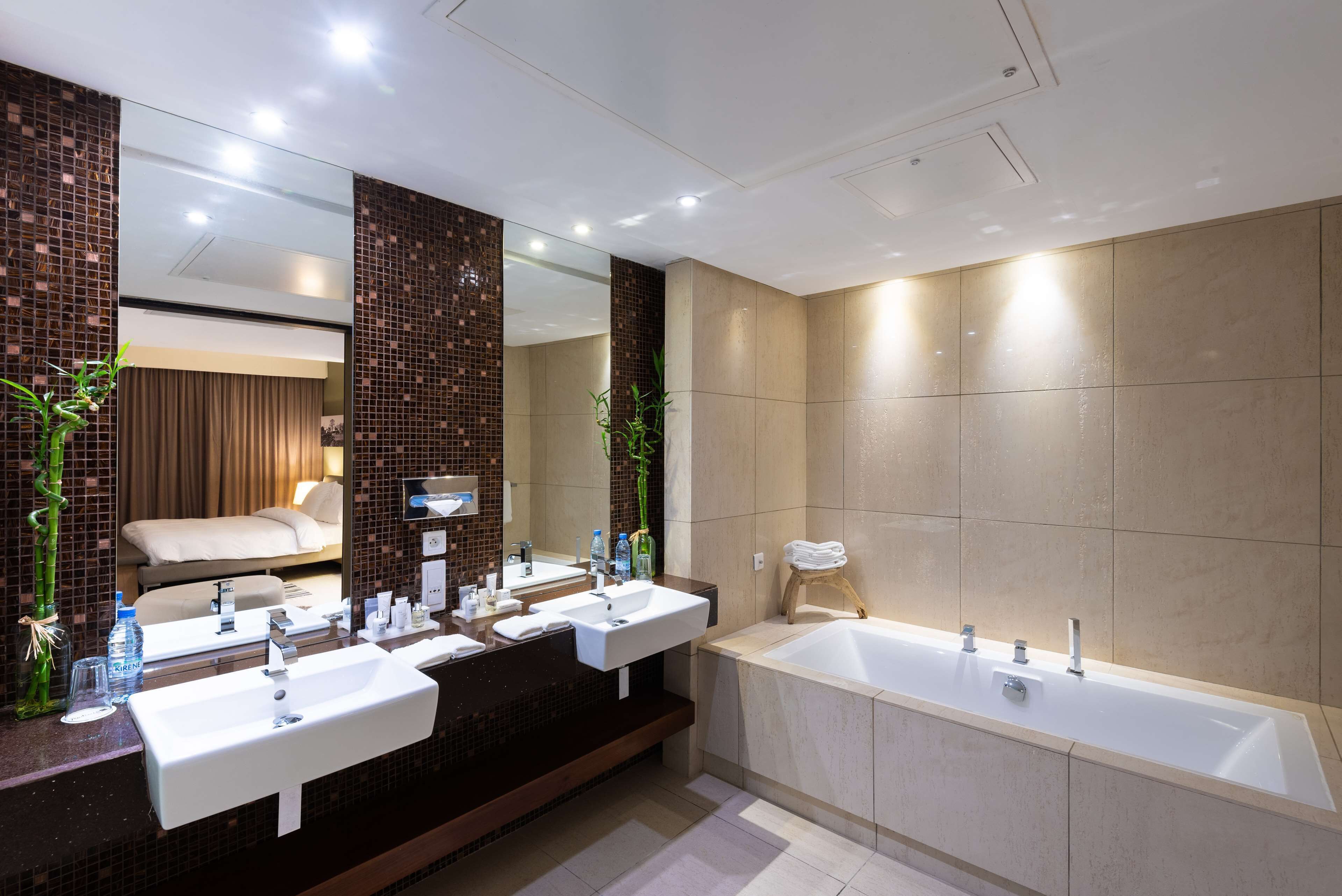 A modern bathroom with two sinks, a large bathtub, and a mirror reflecting a bed.