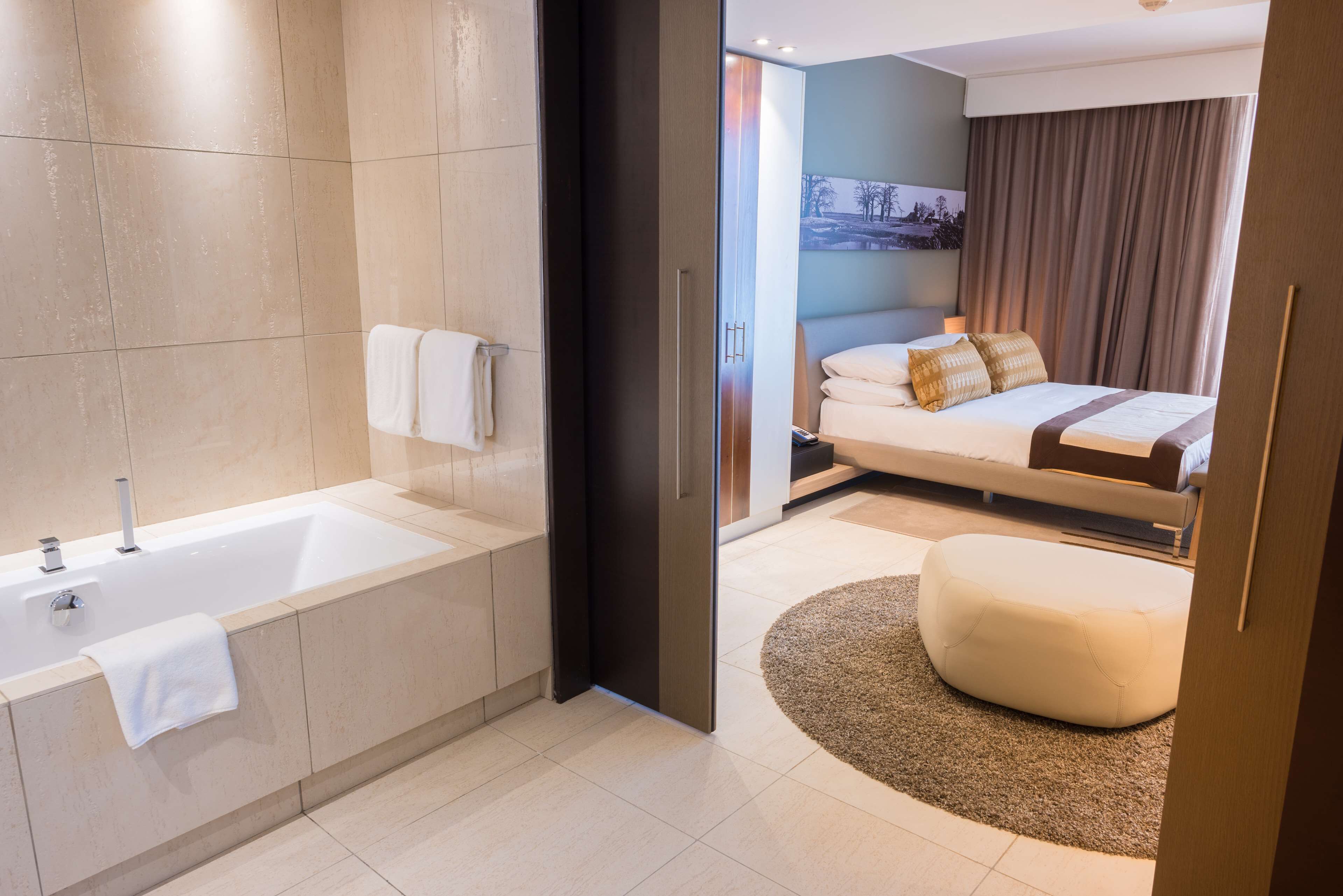 Un baño moderno con bañera, una toalla colgada en la pared, y un dormitorio separado con cama, almohadas y mesita de noche.
