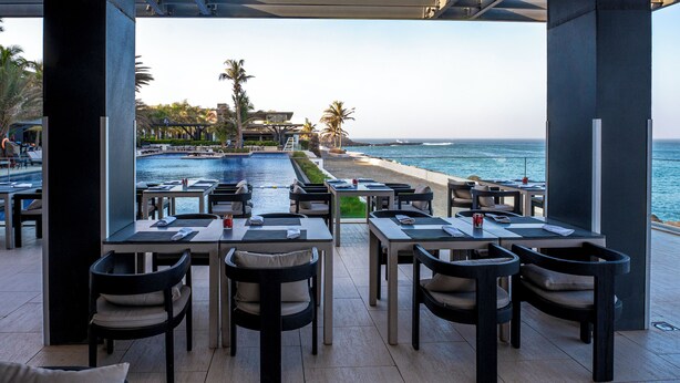 Un área de comedor al aire libre con mesas y sillas, con vista a una piscina y una playa con palmeras y vistas al océano.