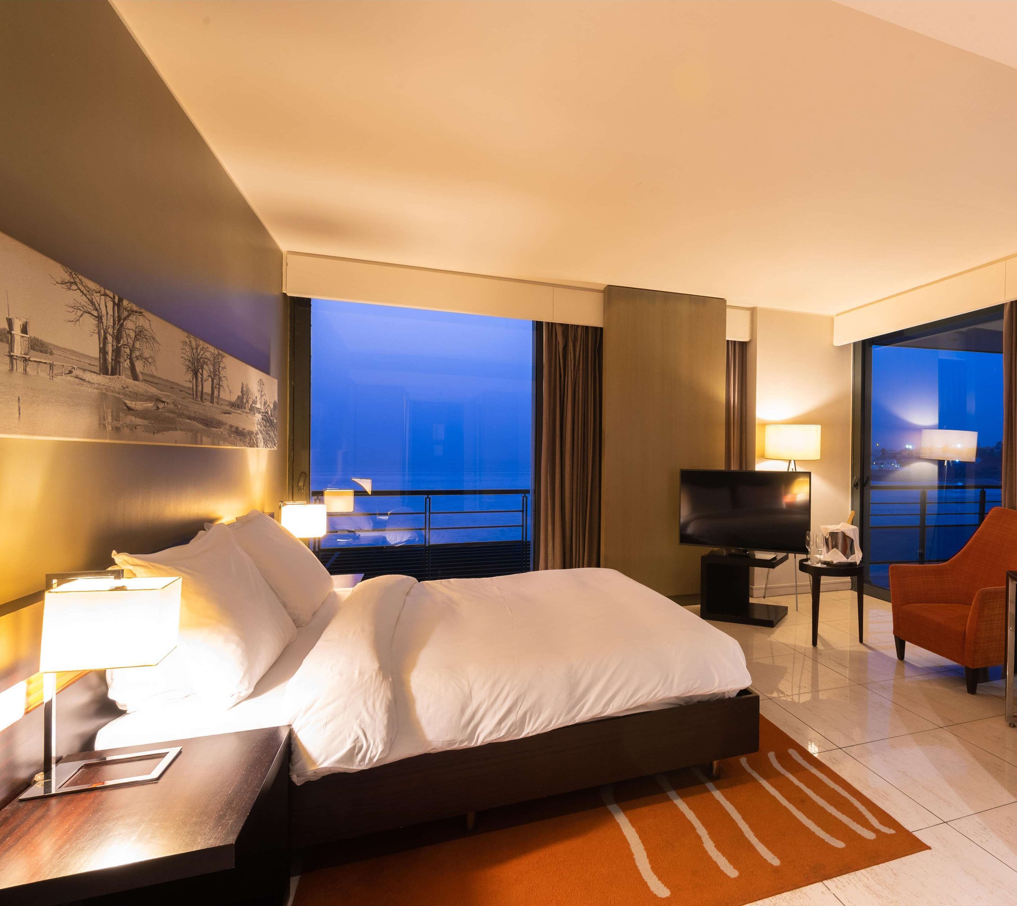 Ein Hotelzimmer mit einem großen Bett, einem Fernseher, einem Schreibtisch mit Lampe und Blick auf die Stadt im Abendlicht.