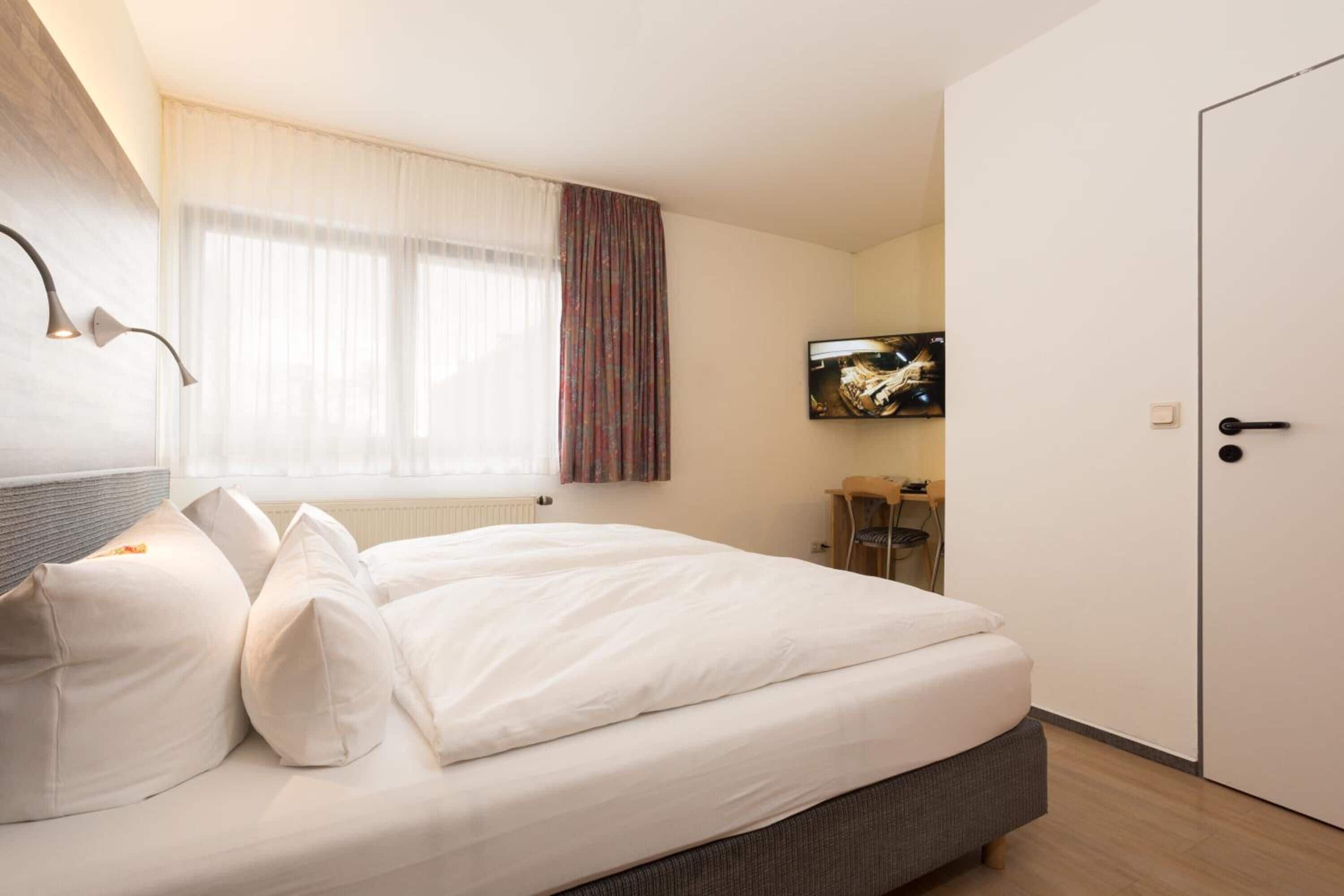 A neatly made bed with white linens and pillows, a television mounted on the wall, and a door to the right.