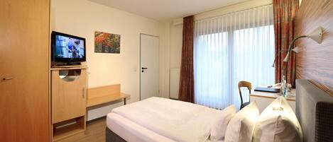 A hotel room with a bed, a TV, a desk, and a window with curtains.