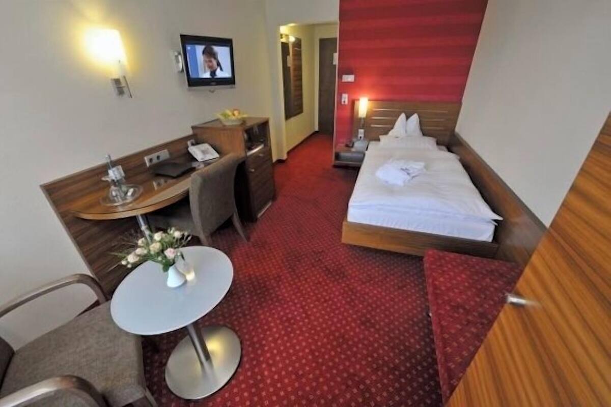 Une chambre d’hôtel avec un lit, un bureau, une chaise, une petite table ornée d’un vase de fleurs, un téléviseur et un sol recouvert d’une moquette rouge.