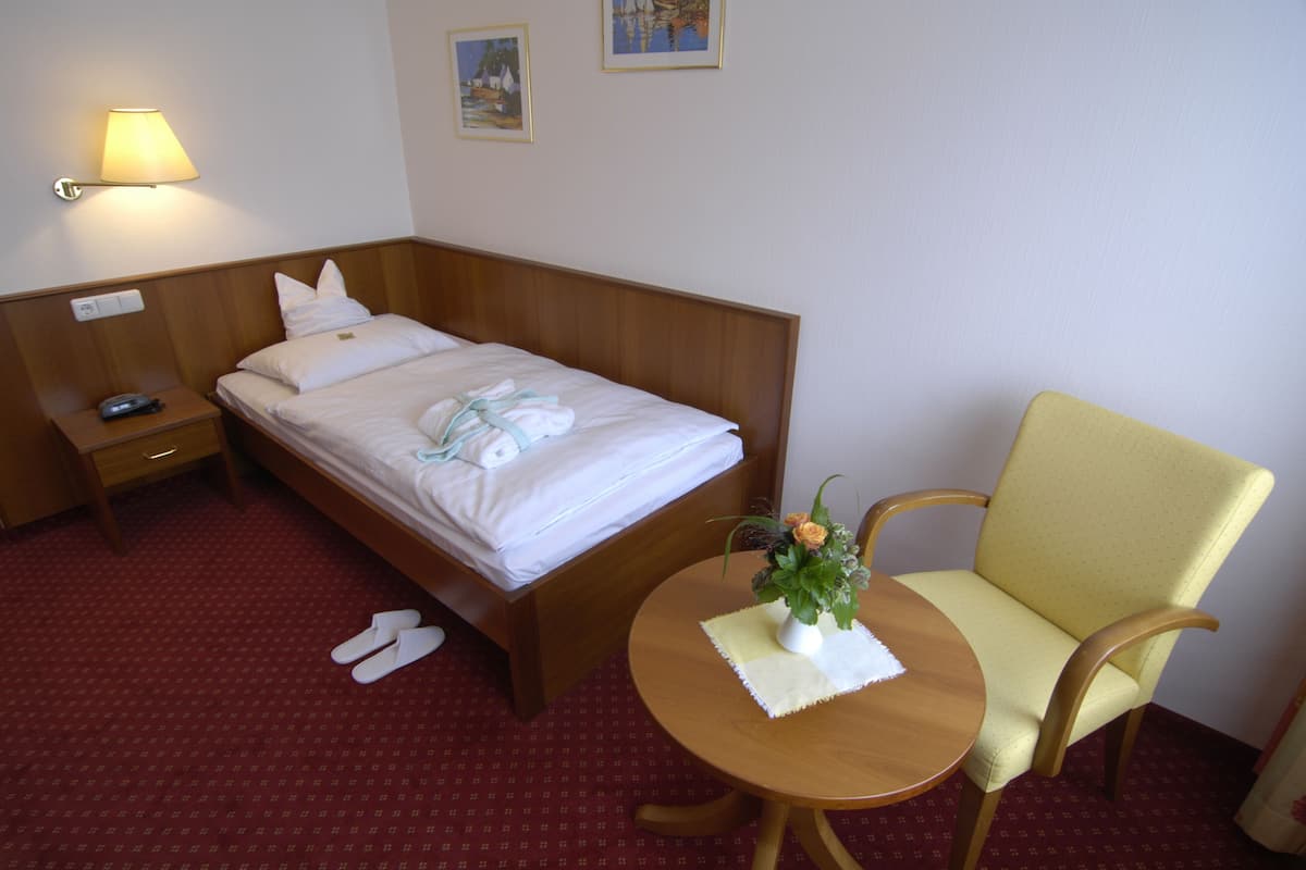 Une chambre d’hôtel avec un lit simple, une table de chevet avec une lampe, une chaise et une petite table sur laquelle est posé un vase de fleurs.