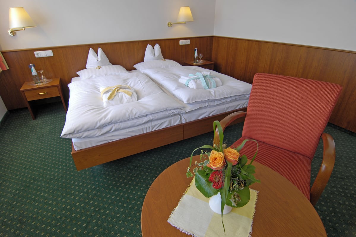 Une chambre d’hôtel avec deux lits, une tête de lit en bois, une chaise rouge et une petite table sur laquelle est posé un vase de fleurs.