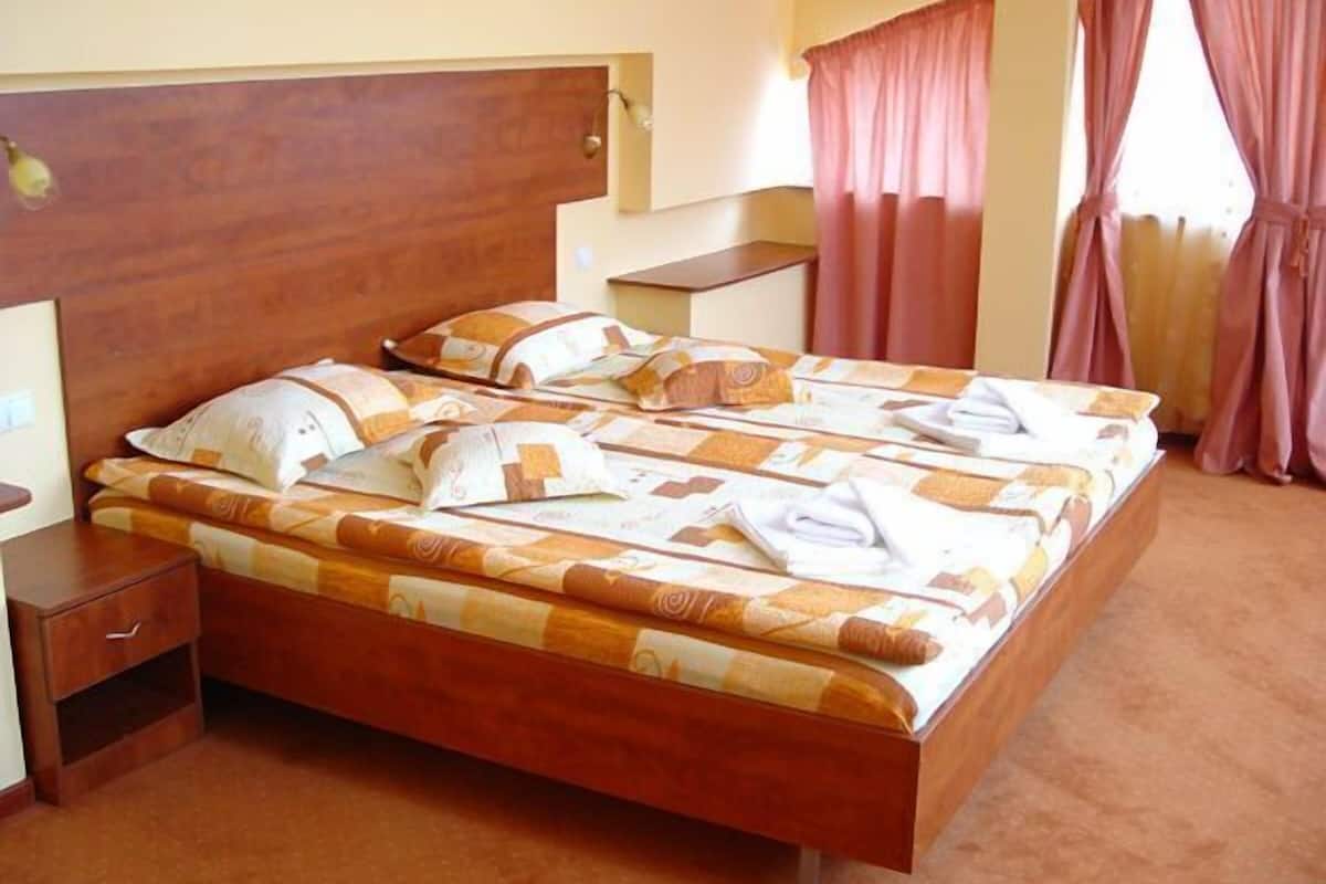Ein Hotelzimmer mit hölzernem Kopfteil, einem Bett mit gemustertem Bettbezug, einem Nachttisch und einem Fenster mit Vorhängen.