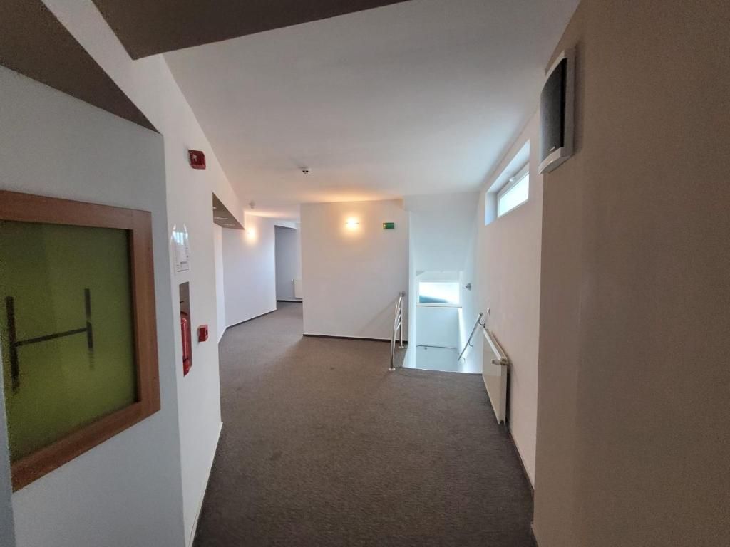 A corridor with a carpeted floor, a door with a green frame, and a window on the right.