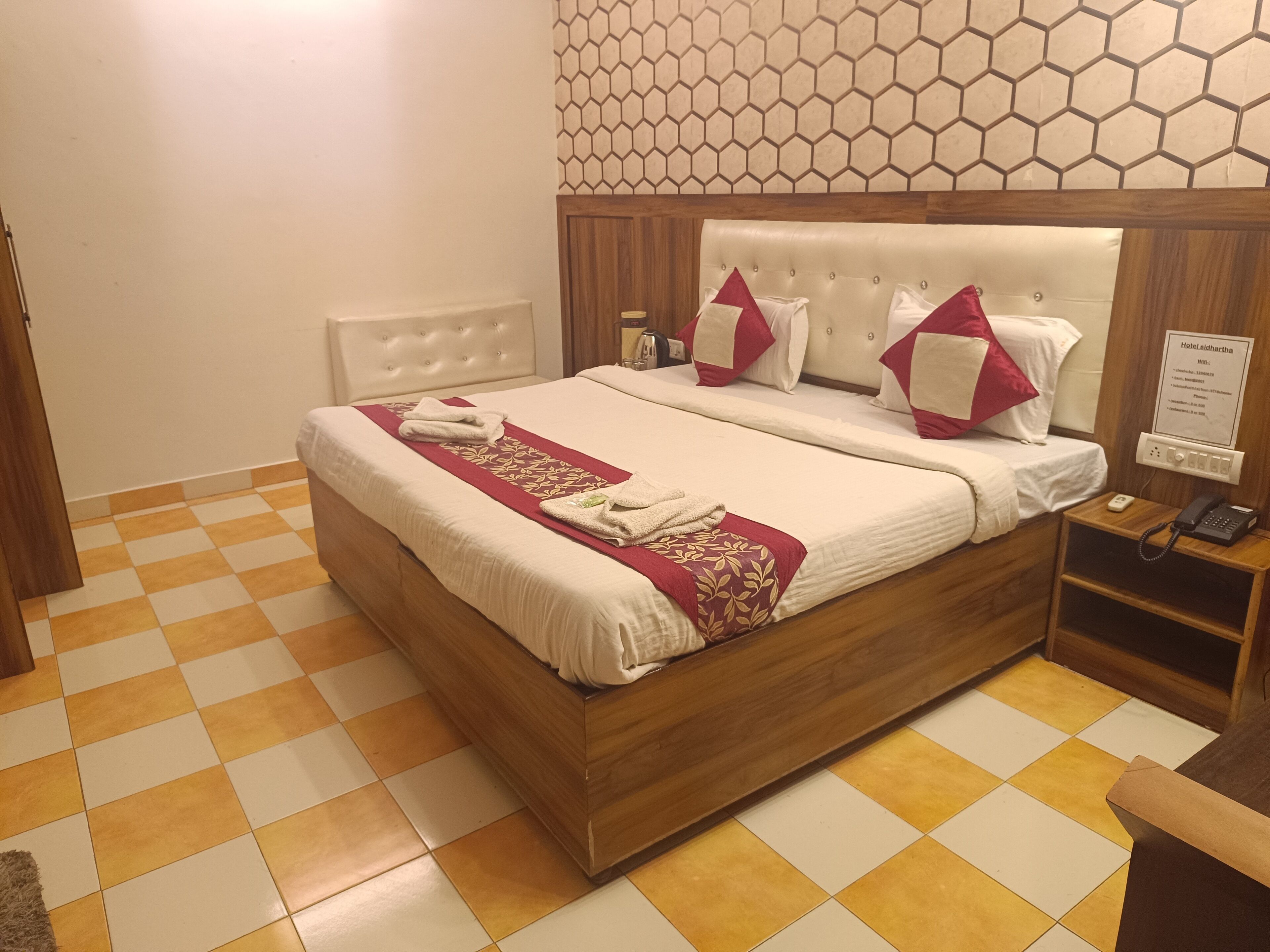 Une chambre d’hôtel avec un sol à carreaux, un lit avec une tête de lit en bois, du linge de lit blanc et un plaid à motifs rouges et blancs.