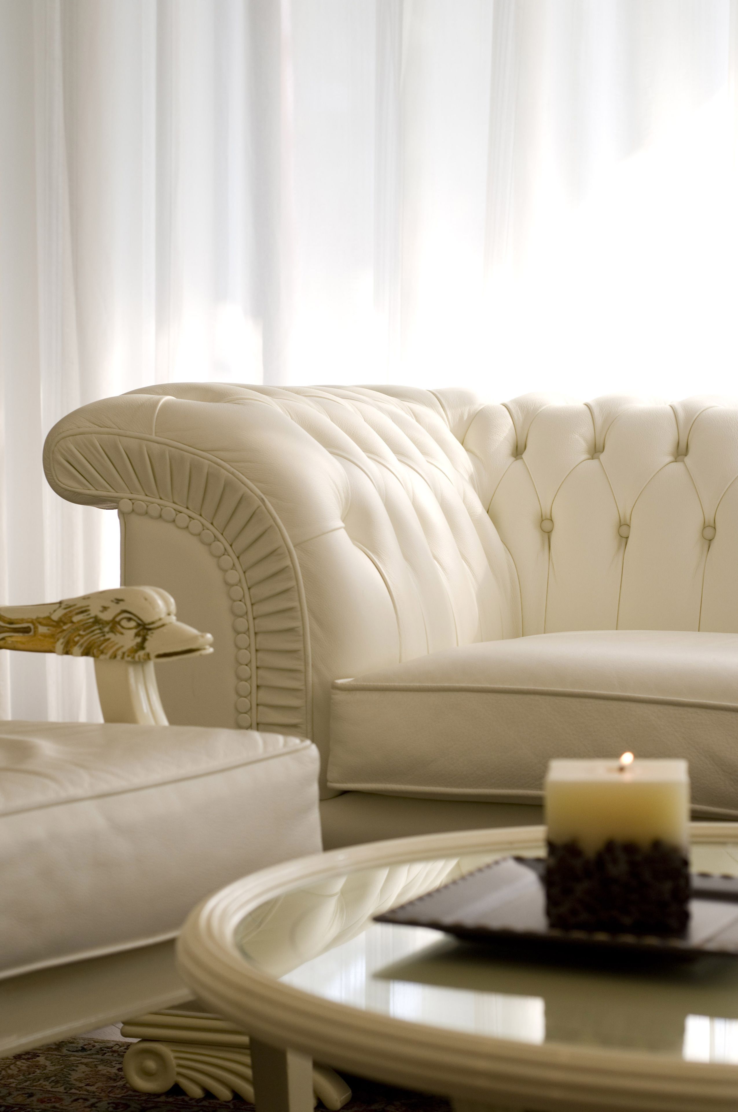 A plush white sofa with a tufted backrest, a round glass coffee table with a candle, and a patterned rug.