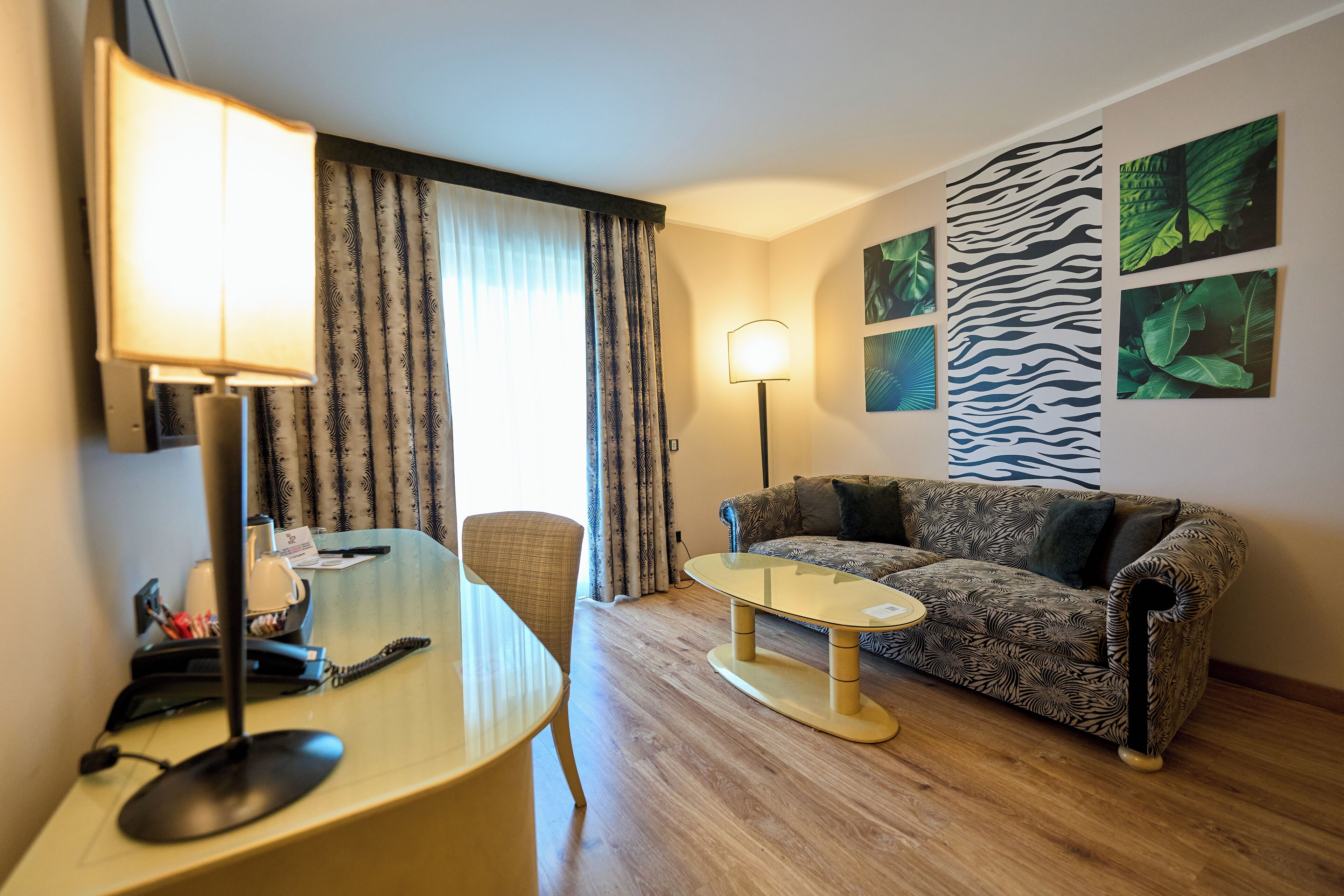 A hotel room with a desk, chair, sofa, and a large window with curtains.