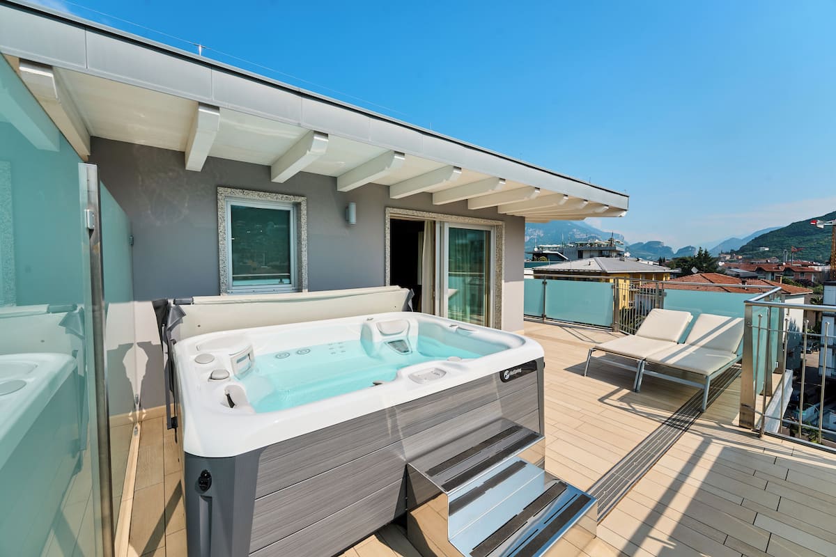 Un jacuzzi en una terraza con cerramiento de vidrio, una zona de estar cubierta y vistas a un pueblo y montañas al fondo.