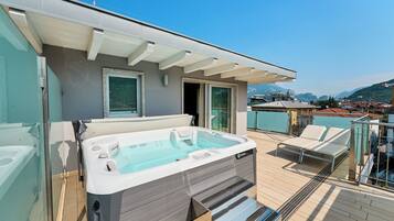 A hot tub on a deck with a glass enclosure, a covered seating area, and a view of a town and mountains in the background.