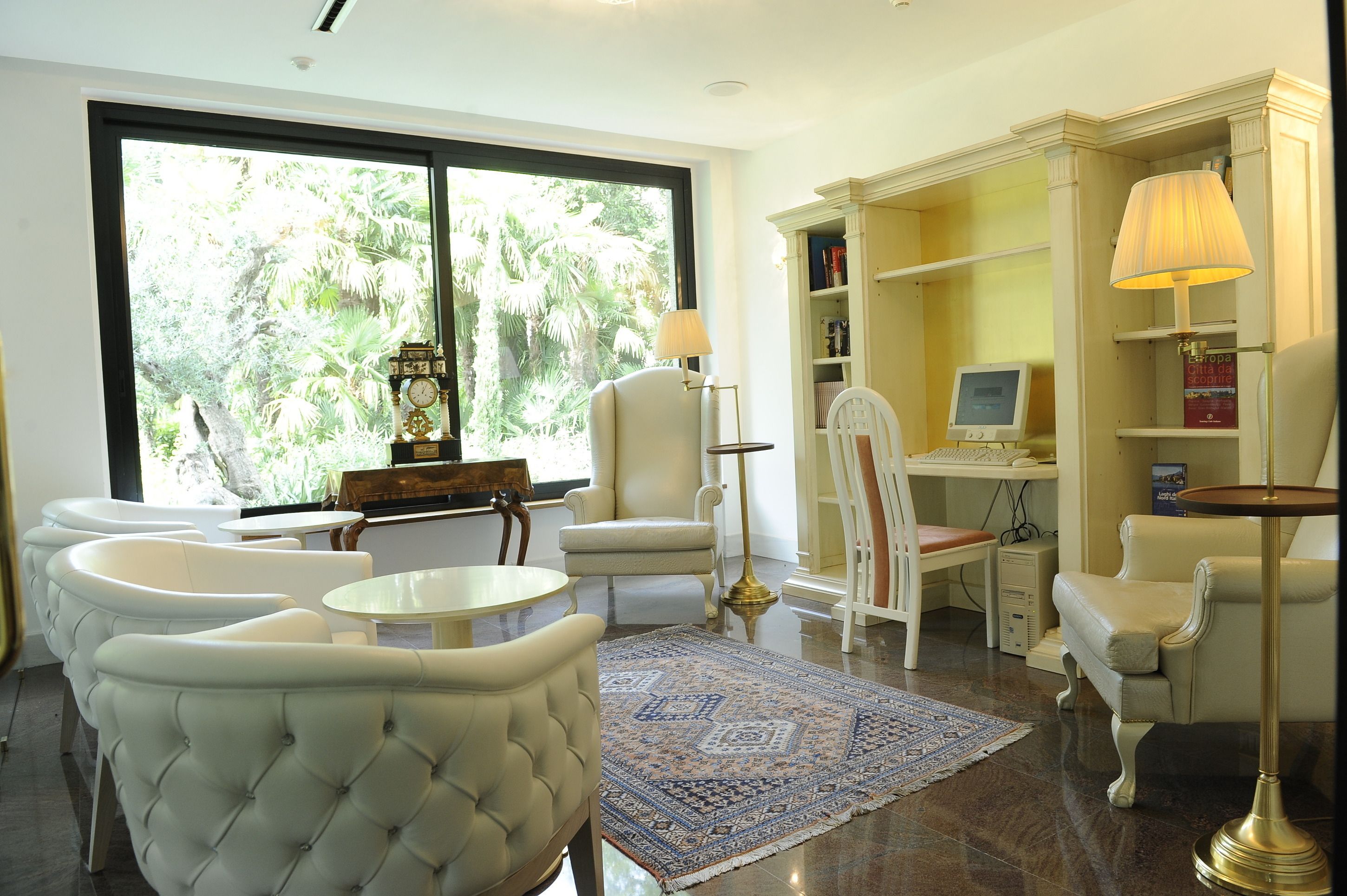 A spacious living room with a large window, a desk with a computer, and a comfortable seating area with chairs and a rug.