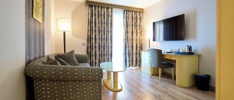 A hotel room with a patterned sofa, a small round table, a desk with a chair, a TV mounted on the wall, and a window with curtains.