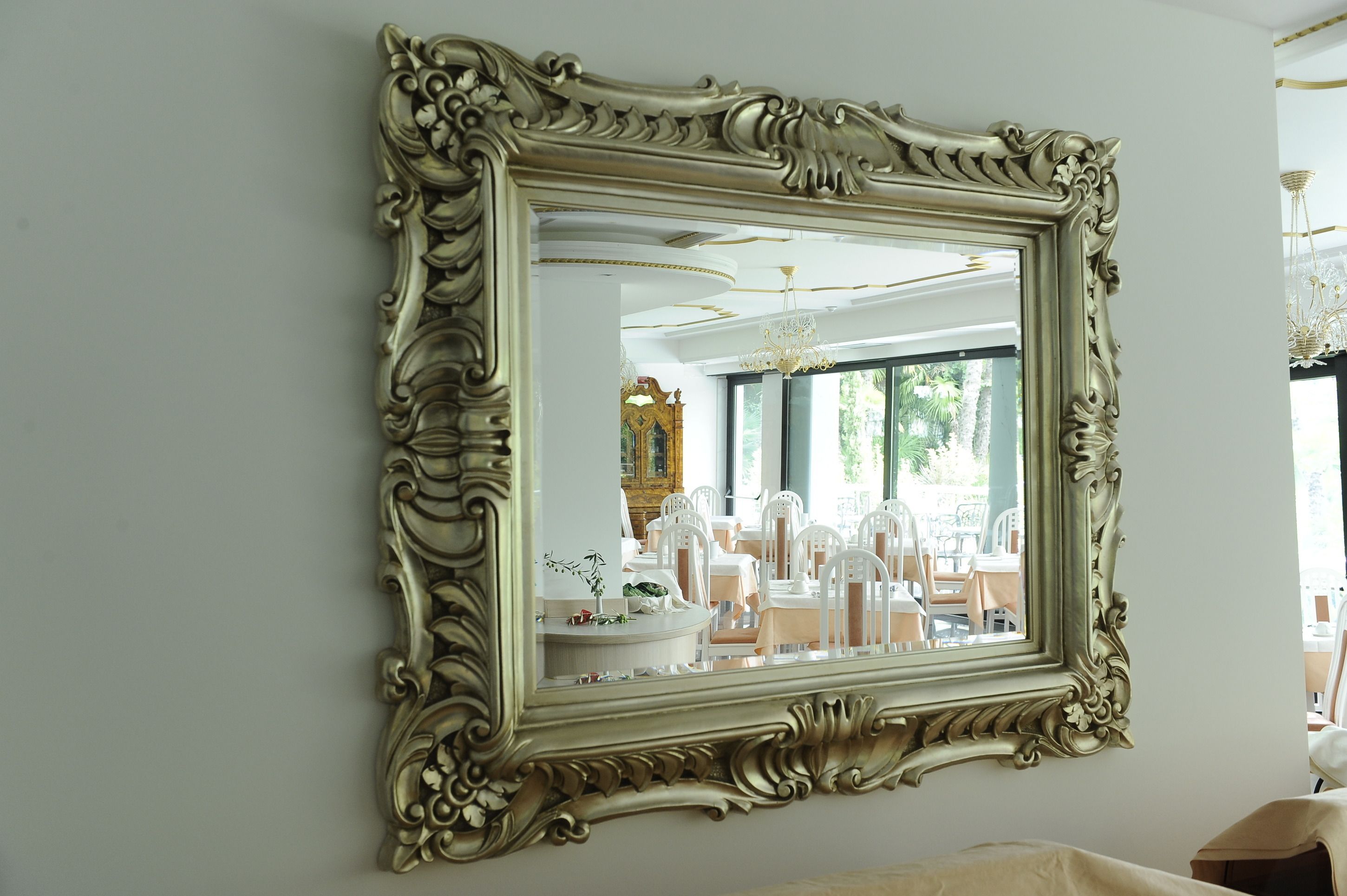A large ornate mirror with a gold frame, reflecting a room with white furniture and a chandelier.