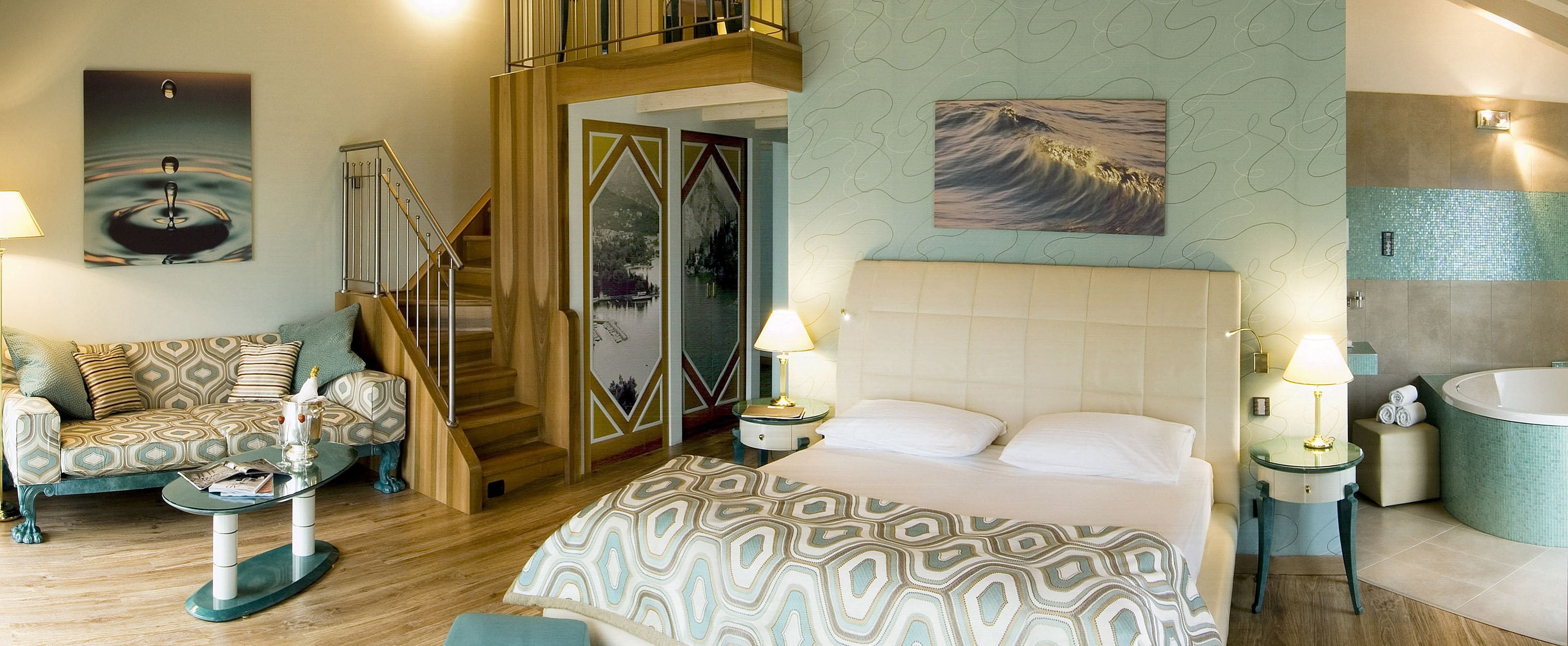 Habitación de hotel con cama, sofá, mesa de centro de vidrio, mesita de noche con lámpara, baño con bañera y escalera.