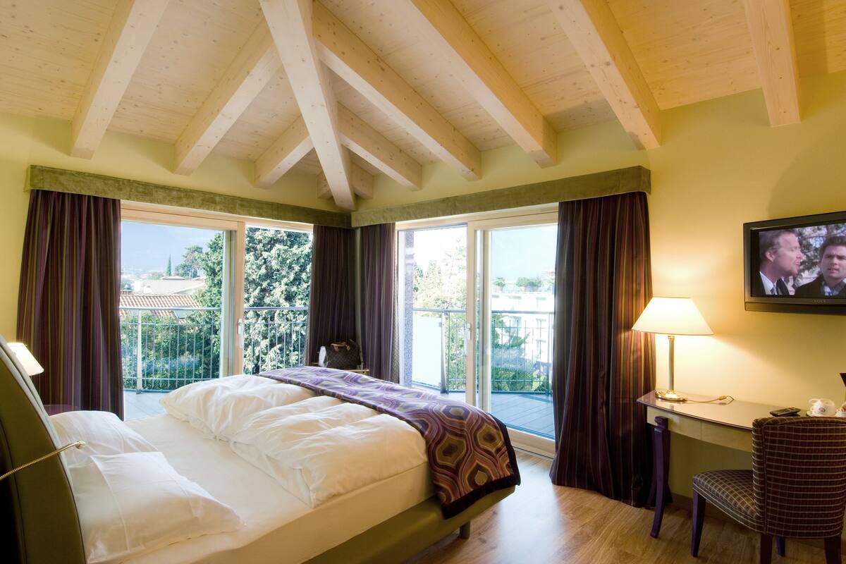 Habitación de hotel con cama, escritorio, silla, televisor y ventanas amplias con cortinas.