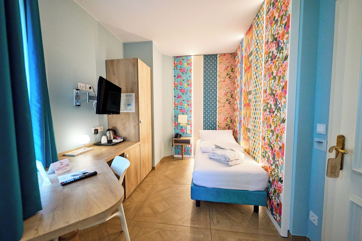 Una habitación de hotel con un escritorio de madera, una cama, un televisor montado en la pared y un papel tapiz floral colorido.