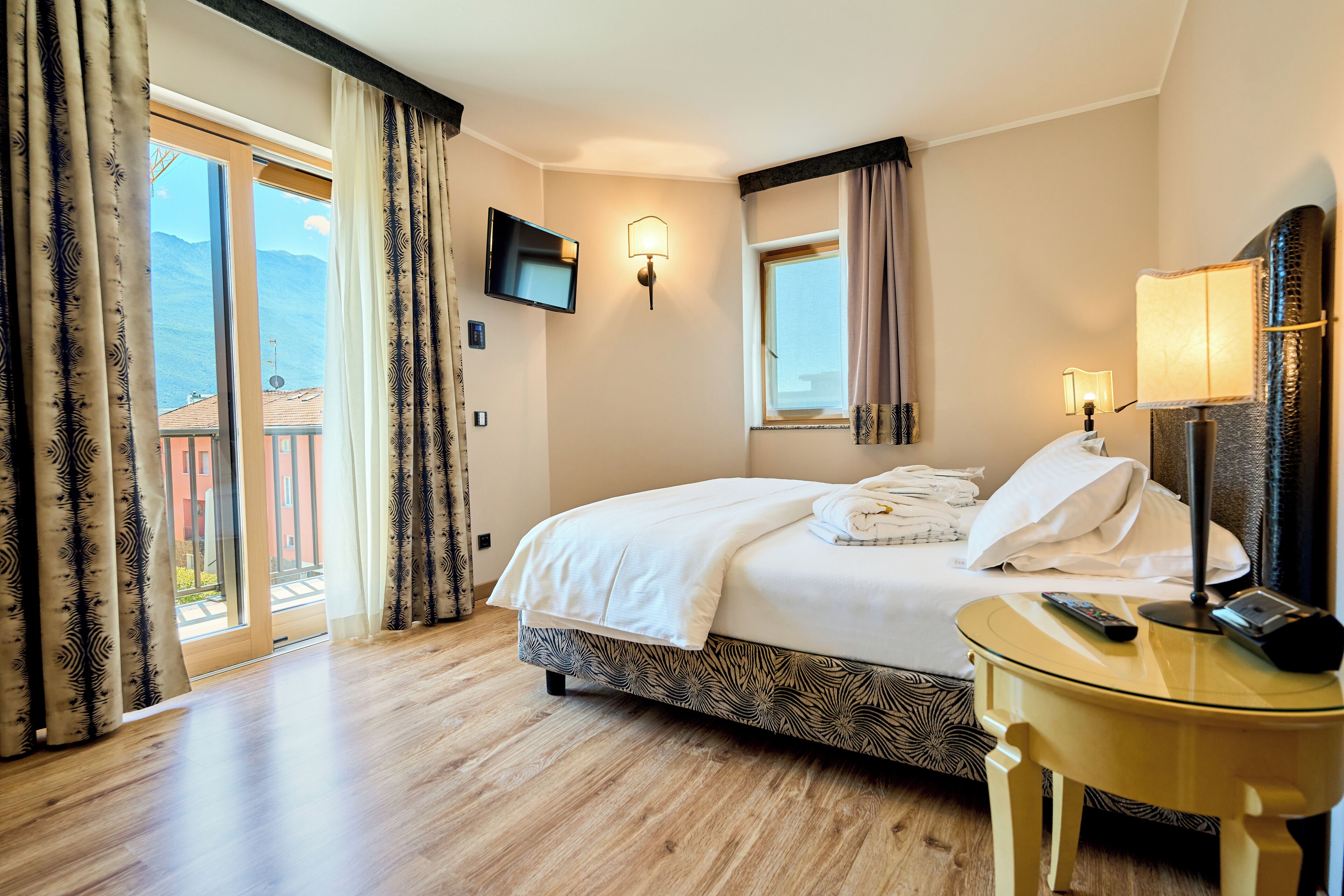 A hotel room with a bed, a TV, a lamp, a table, and a view of mountains through the window.