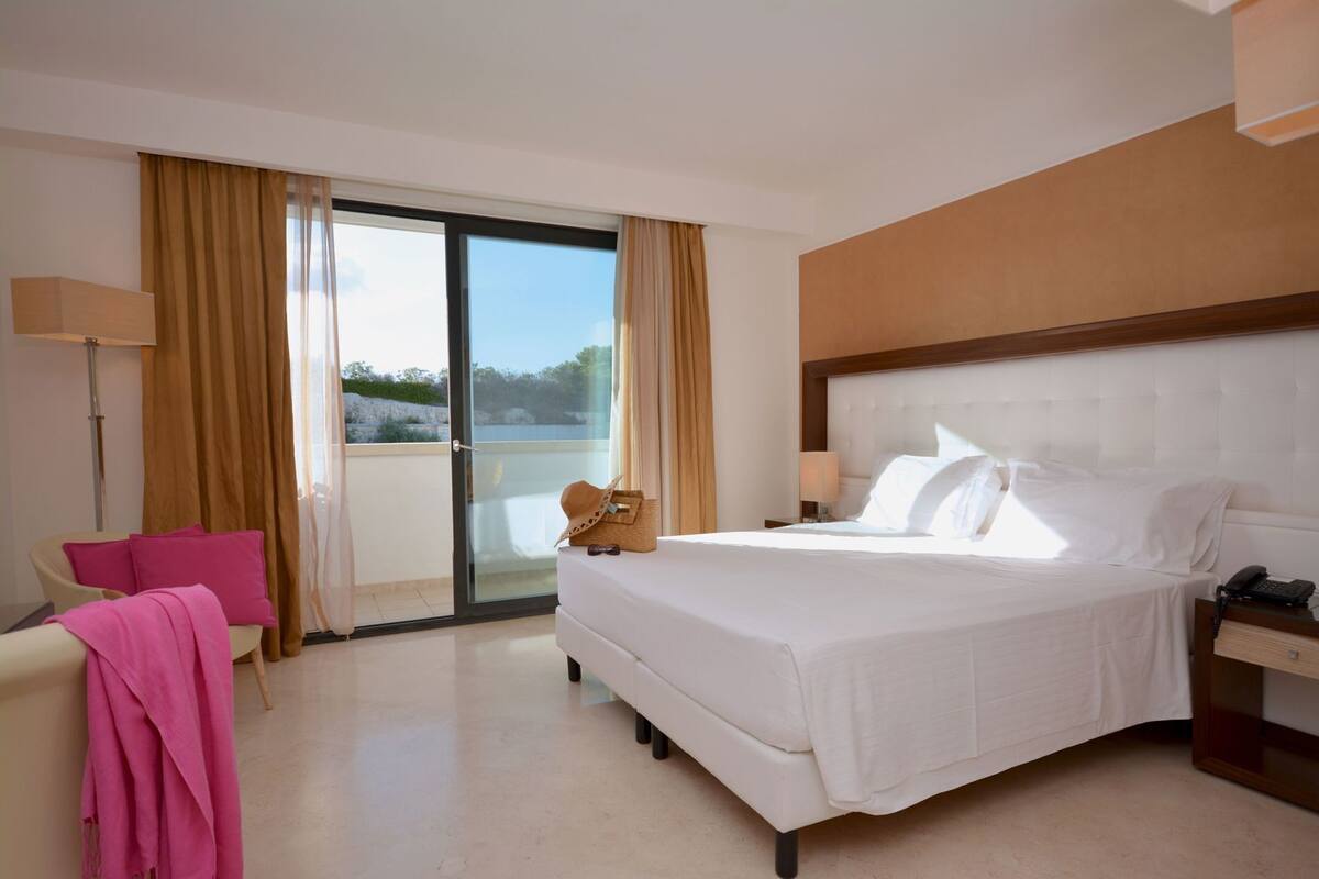 Une chambre d’hôtel avec un lit, une chaise, un bureau, une lampe et une vue sur l’extérieur, visible à travers une porte vitrée.