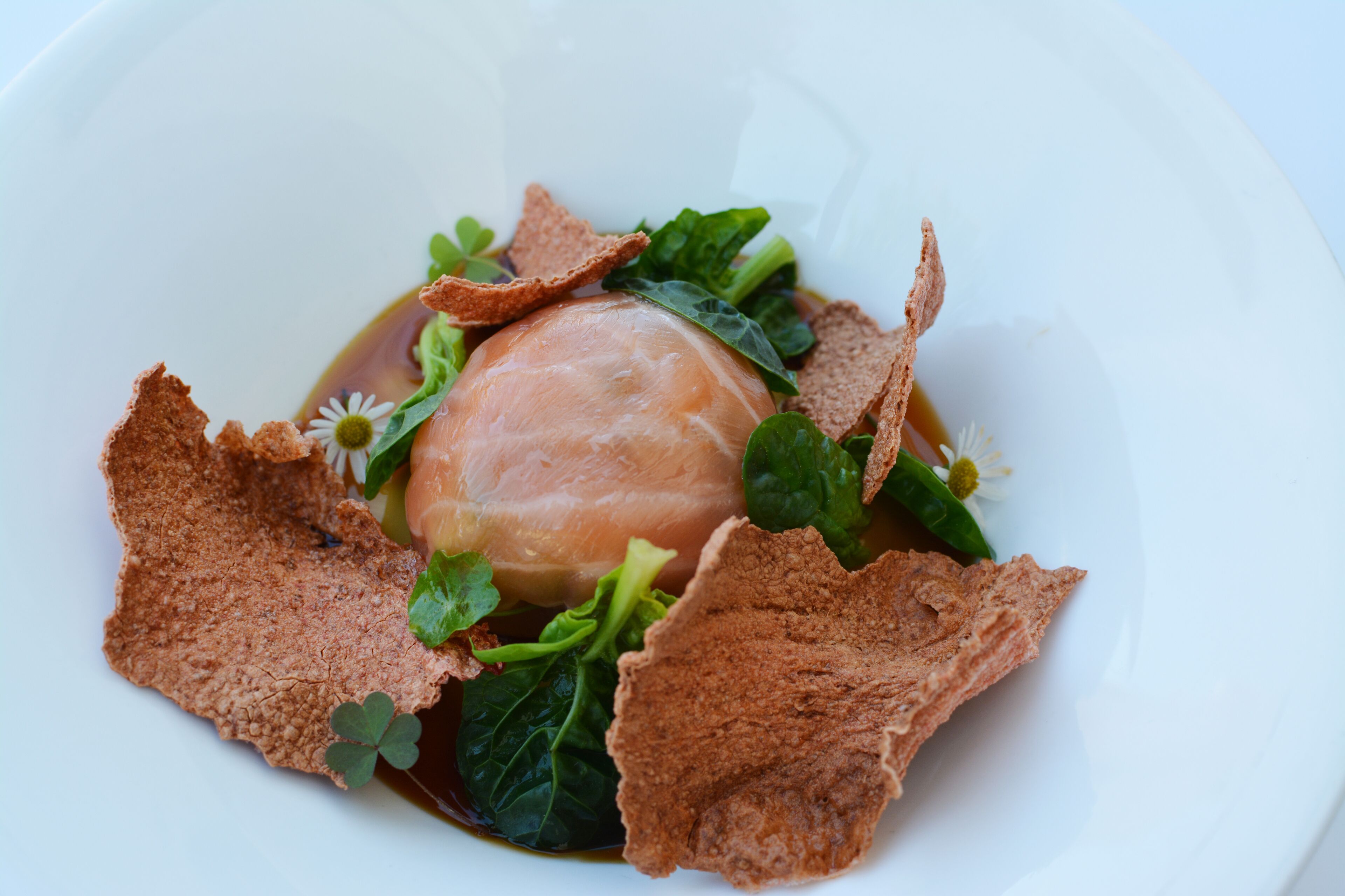 A white plate with a central piece of food, surrounded by greens and brown elements.