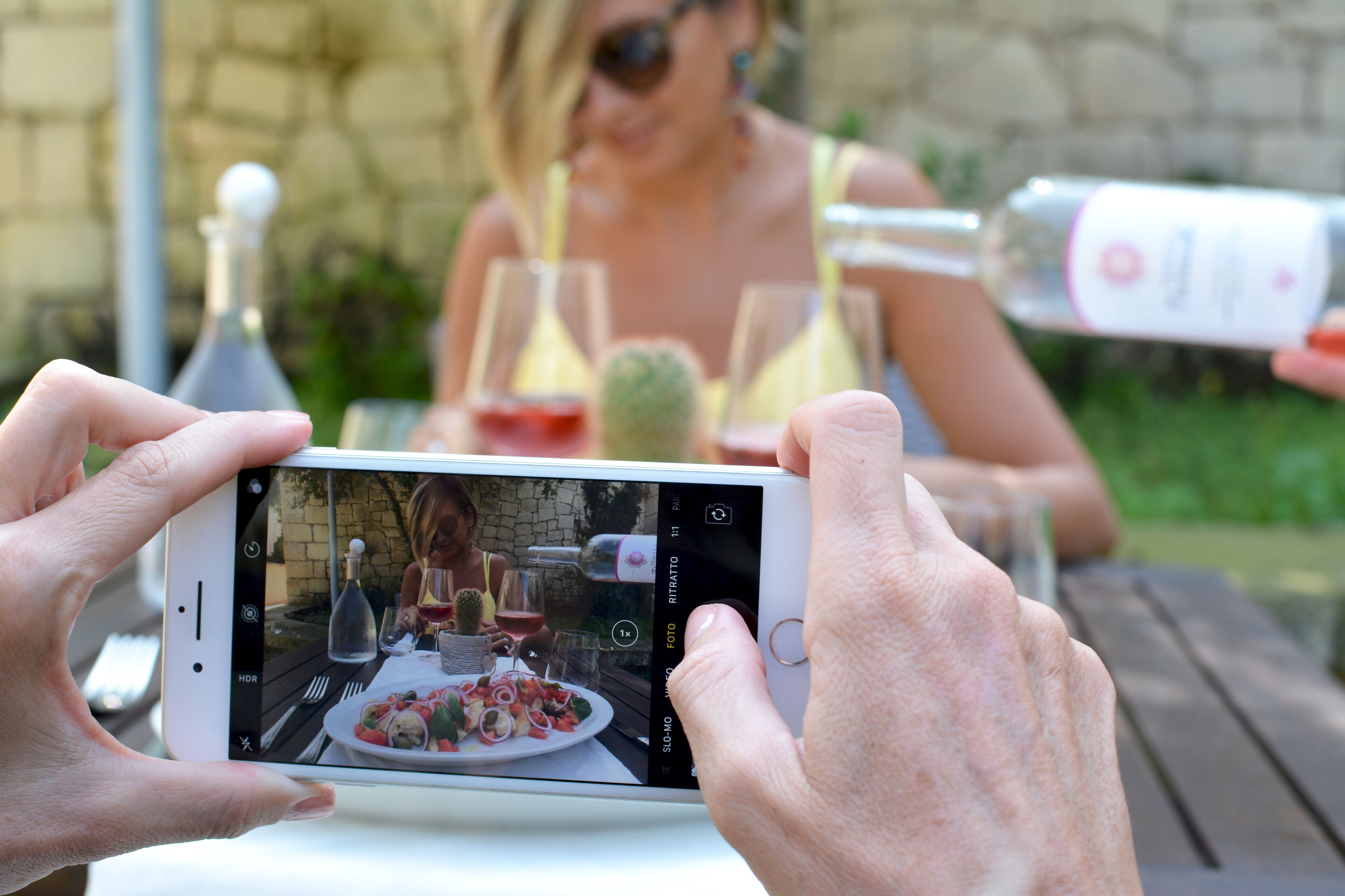 Una persona sosteniendo un teléfono inteligente, grabando una escena con una mesa dispuesta para una comida, una botella de vino y una copa.
