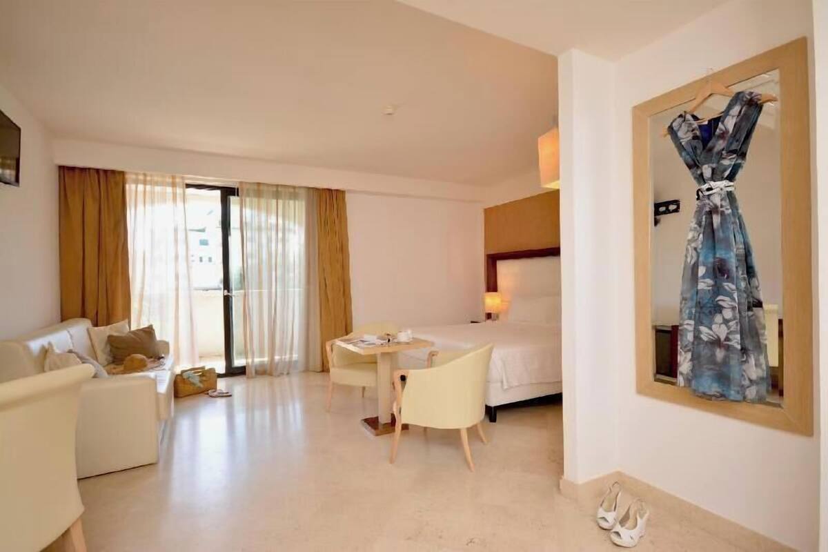 Une chambre d’hôtel avec un lit, un bureau, une chaise, un canapé, un miroir et une paire de chaussures posées sur le sol.