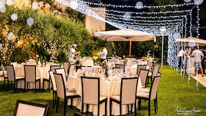 Un área de comedor al aire libre con mesas y sillas redondas, luces colgantes y un fondo de césped.