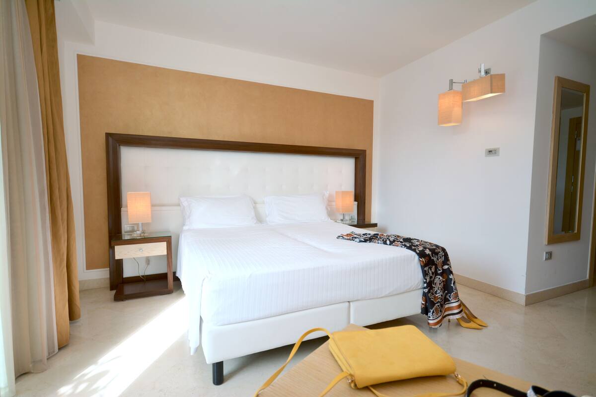 Une chambre d’hôtel moderne avec un grand lit, des lampes de chevet, une table de chevet et un sac jaune posé sur le sol.