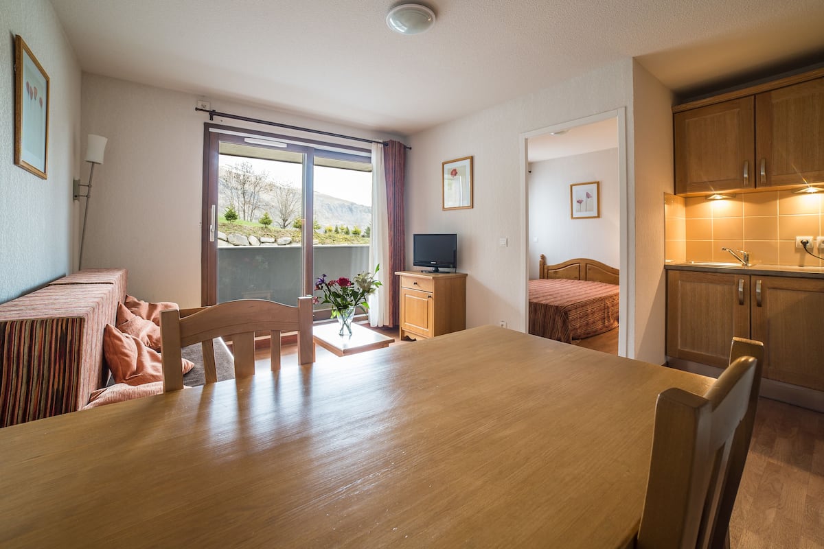 A compact hotel room with a wooden dining table, a single bed, a small kitchenette, and a sliding glass door leading to a balcony with a view of greenery.