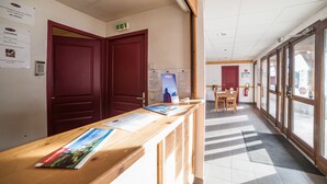 A reception area with a wooden counter, red doors, informational posters, and a view through large windows. - Noemys les Terrasses de la Toussuire  (Saint-Pancrace)