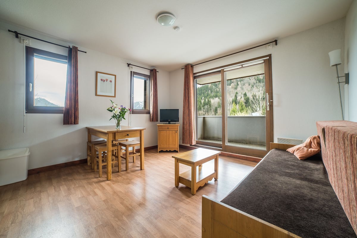 A room with a wooden floor, a sofa, a TV, a table, and a window with curtains.