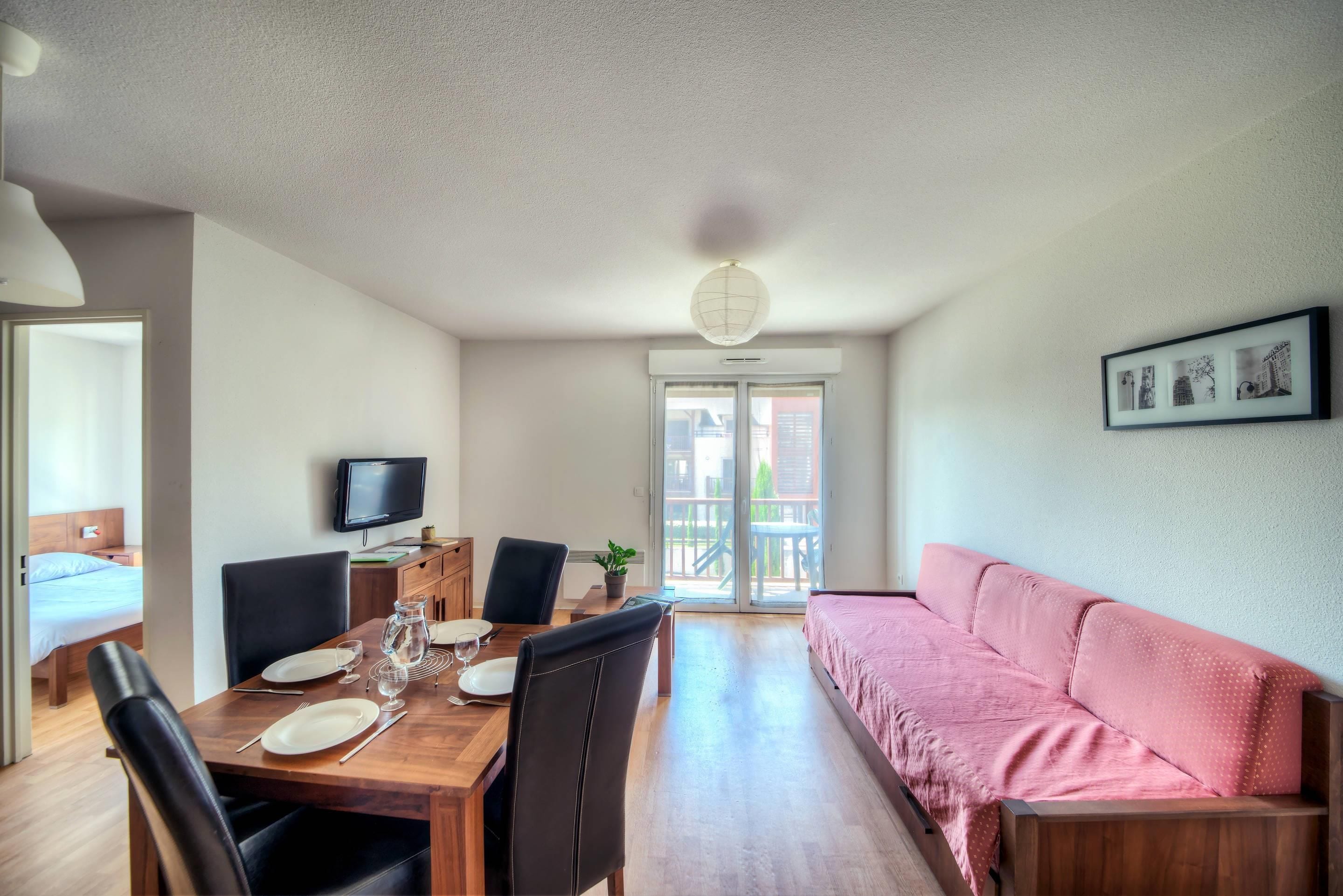Un comedor con una mesa de madera dispuesta para dos personas, un televisor encima y un balcón con vistas.