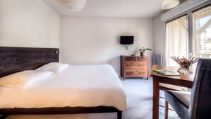 A hotel room with a large bed, a wooden dresser, a desk with a chair, a flat-screen TV mounted on the wall, and a window with curtains.